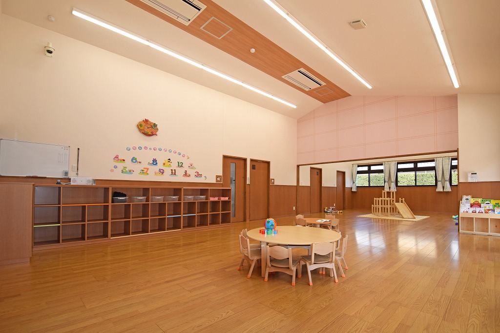 にこにこ保育園の保育室竣工写真。勾配屋根を活かした開放的な高天井と、家庭的な温もりを感じるミディアムブラウンを基調とした落ち着いた内装デザイン