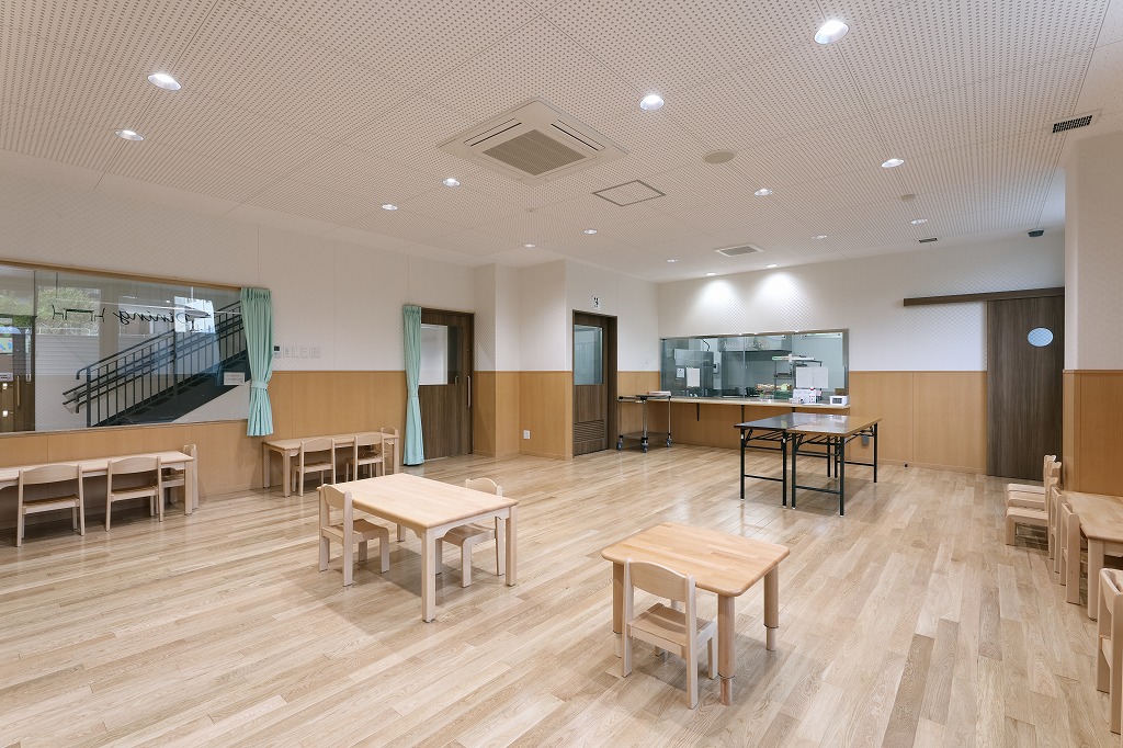 厨房が見える大きなガラス窓を配した、木製テーブルと椅子が並ぶ真愛三ツ谷こども園の明るいランチルーム(共同食堂)